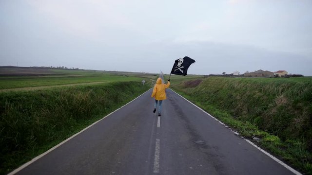 A young girl with a yellow raincoat runs along a road between fields heading to the coast waving a pirate flag.
