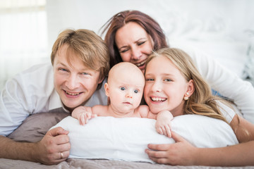 Fototapeta premium Beautiful smiling young family mom dad elder daughter and newborn baby are lying on a large bed in a bright bedroom. Concept of friendly caucasian family