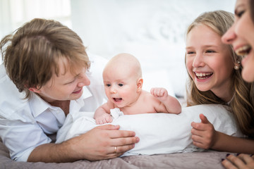 Beautiful smiling young family mom dad elder daughter and newborn baby are lying on a large bed in a bright bedroom. Concept of friendly caucasian family