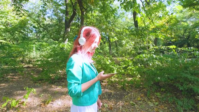 Young happy woman in colorful suit and with pink hair walk in summer park and listen to music
