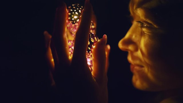 Magic hot light from lanterns falls on the woman eyes. The naked adult sexy caucasian girl lies at night on the bed and holds warm lamps in her hands. Christmas toy in black background. Indoor.