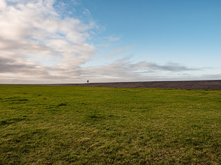 Fototapeta na wymiar Norddeich, Germany. 7 December 2019. Single person walking on the dyke in the distance behind a green field on a sunny winter day.