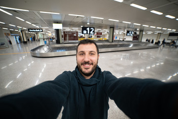 happy man passenger take selfie photo in airport at baggage claim area waiting for his bag trolley