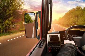Truck dashboard with driver's hand on the steering wheel and side rear-view mirror on the countryside road against sky with sunset - 309848879