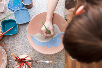 Potter woman paints a ceramic plate. Girl draws with a brush on earthenware. Process of creating clay products. 