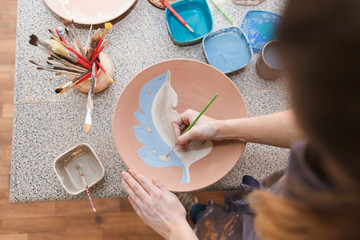 Potter woman paints a ceramic plate. Girl draws with a brush on earthenware. Process of creating...