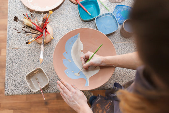 Potter woman paints a ceramic plate. Girl draws with a brush on earthenware. Process of creating clay products. 