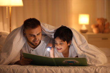 Father and son with flashlight reading book under blanket at home