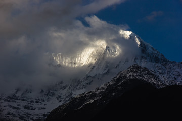 Plakat A view of the summit of the Himalayan peak of Annapurna South lit by the sun as clouds hover around it as seen from the Annapurna Base Camp trail.