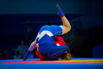Two woman in blue and red wrestling on a yellow wrestling carpet in the gym..