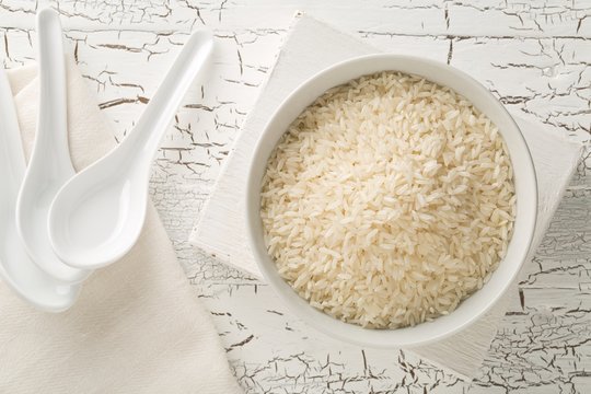 White uncooked, raw long grain rice in white bowl with spoons on rustic white wooden table top view flat lay from above