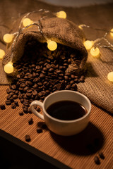 A cup of coffee and a jute bag with grains of non-ground coffee. Beautiful still life. The lights of a garland in the background.