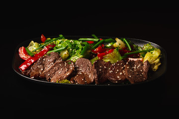 Juicy Beef Sirloin Steak Salad with bell pepper, sesam seeds and green vegetables in a black plate, on black background. Healthy food