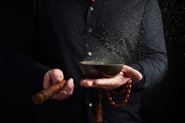 adult man in a black shirt rotates a wooden stick around a copper Tibetan bowl with water