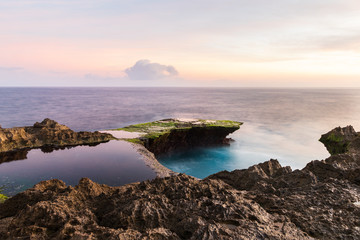 Fototapeta na wymiar Devil's Tear at sunset, island of Nusa Lembongan, Bali, Indonesia. Rocky shore in foreground. Yellow sky with clouds beyond. 