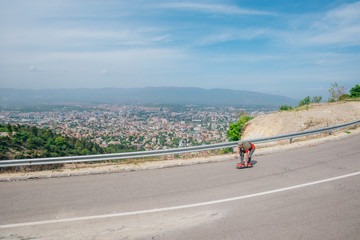 Male caucasian longboarder riding downhill on an empty road doing a speed tuck and grabbing the board while driving the longboard fast. Wearing a red t-shirt green hat and black jeans.