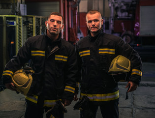 Portrait of two young firemen in uniform standing inside the fire station .