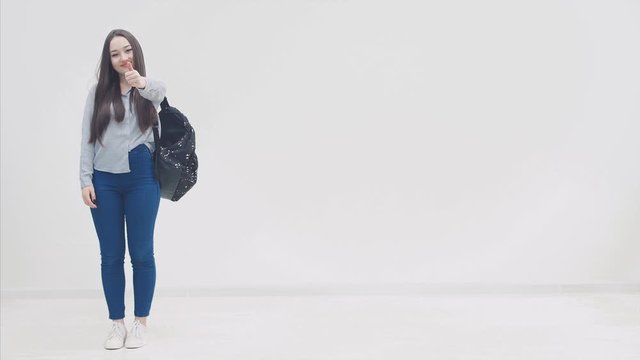 Happy, smiling, full height confident girl with long hair appearing on a white background with backpack behind her shoulders, giving thumbs up.