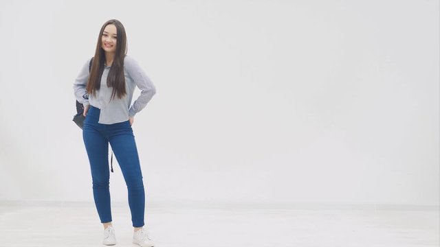 Happy, smiling, full height asian girl with long hair appearing on a white background with backpack slung over her shoulder, hands in back pockets of her jeans, posing.