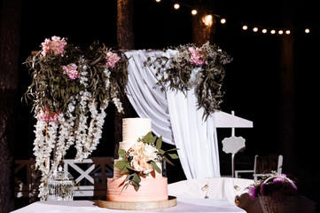 Beautiful four tiered wedding cake decorated with a flowers, outdoors near the festive arch, in the evening