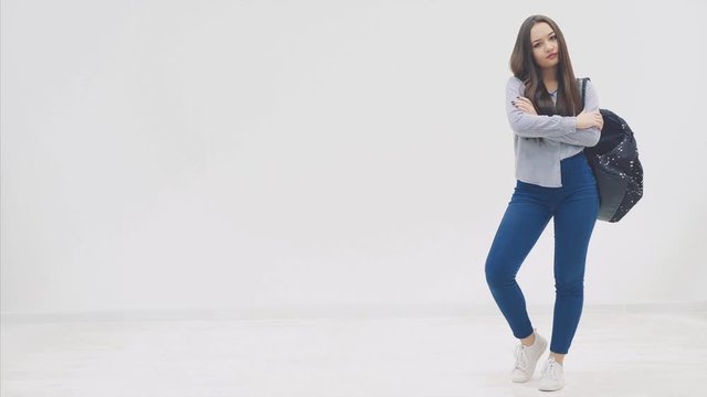 Full height asian, girl with long hair appearing on a white background with backpack slung over her shoulder, and hands folded. Sour face expression.
