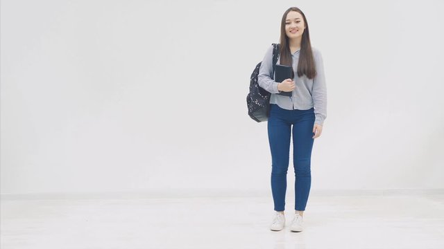 An asian schoolgirl appearing on a white background. With a backpack. Holds the book and smiles.