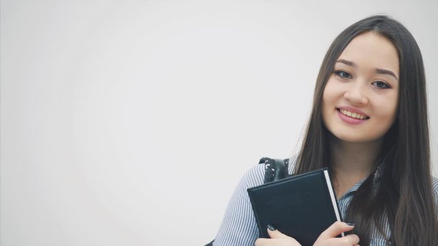 An asian schoolgirl appearing on a white background with a backpack slung over her shoulder, holding book near chest, posing and smiling.