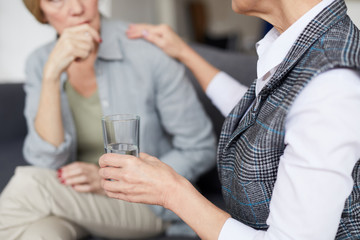 Cropped image of female psychologist comforting patient during therapy session and offering glass of water, copy space
