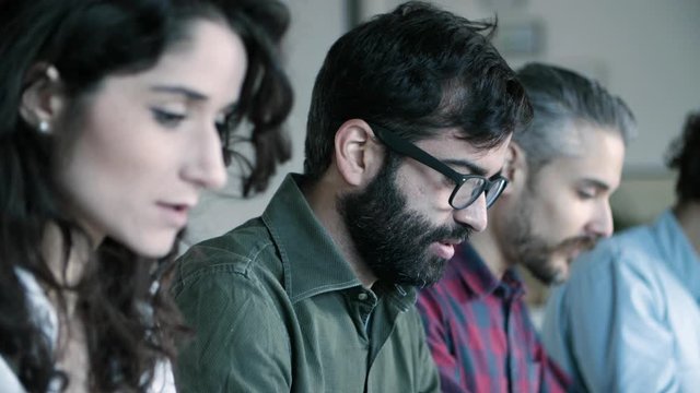 Side view of cheerful developer in eyeglasses working with laptop. Young people sitting in row and typing on laptops. Technology concept