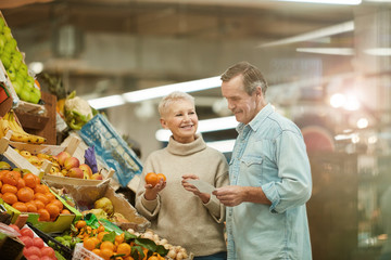 Waist up portrait of smiling senior couple standing at fruit stand while enjoying grocery shopping in farmers market, copy space