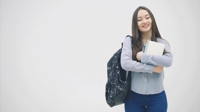 An asian schoolgirl appearing on a white background with a backpack slung over her shoulder, holding a stack of books near chest, posing, smiling.