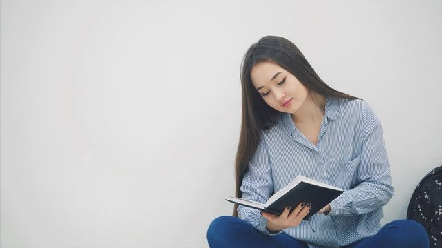 Young asian girl involved in the process of reading book, closing itand hugging, while sitting on the floor in lotos position.