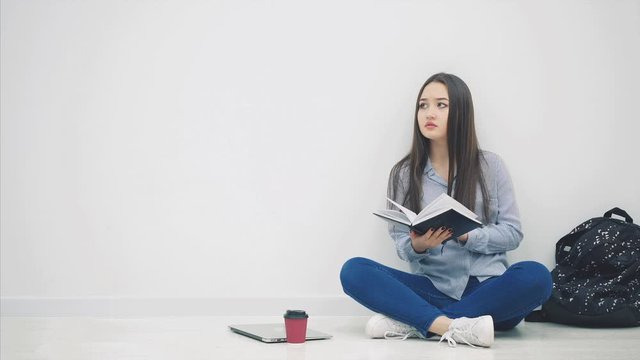Young asian girl involved in the process of reading book and looking around while sitting on the floor in lotos position.