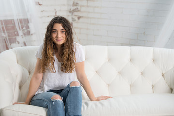 Beautiful young brunette woman relies on white couch wearing white t-shirt and blue jeans