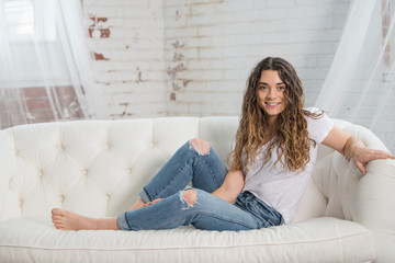 Obraz premium Beautiful young brunette woman relies on white couch wearing white t-shirt and blue jeans