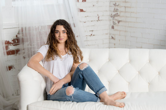 Beautiful young brunette woman relies on white couch wearing white t-shirt and blue jeans