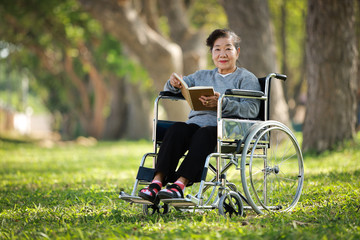 Asian senior woman sitting on the wheelchair and reading book in the park garden smile and happy...