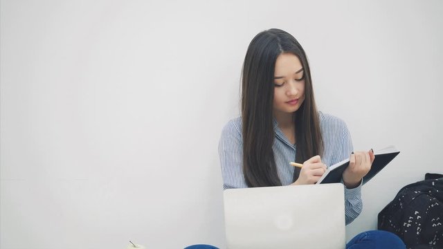 Asian lady sitting on the floor in lotos position, with laptop, copying something into her notebook.