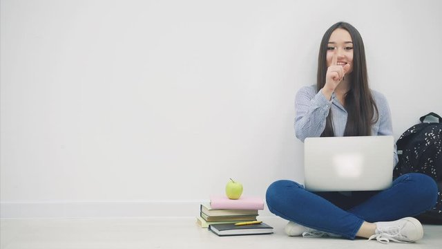 Lovely young asian lady sitting on the floor in lotos position, with laptop, pointing her fingers at the camera, then to the side, smiling.