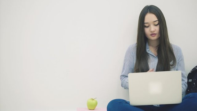 Tired young asian lady sitting on the floor in lotos position, with laptop, yawning, making a break, eating apple.
