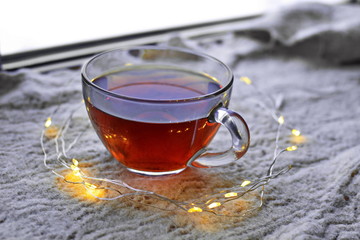 Glass Cup of evening hot tea in circle by glowing garland of yellow lights stands on surface wool white knitted blanket