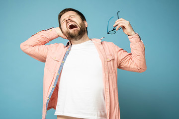 Bearded man yawns funny and stretches, holding glasses in his hands. Isolated on a blue background.