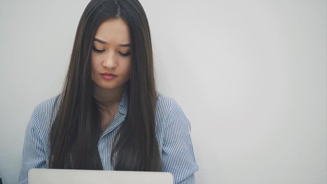 Lovely but tired asian girl sitting with laptop on her knees, having some difficulties, shaking her head, readjusting hair, looking upset.