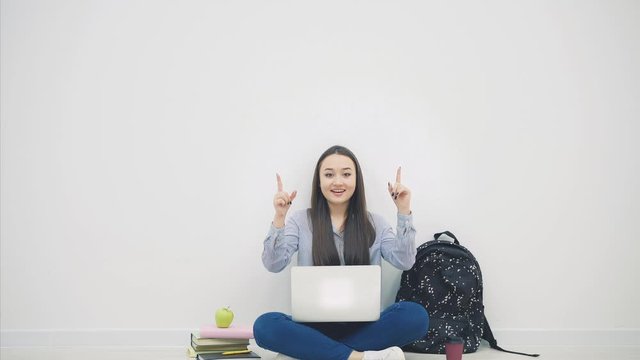 Lovely smiling asian student sitting on the floor in lotos position, with laptop, pointing her fingers up, smiling.