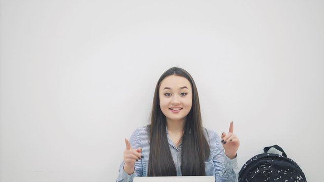 Lovely smiling asian student sitting on the floor in lotos position, with laptop, pointing her fingers up, smiling.