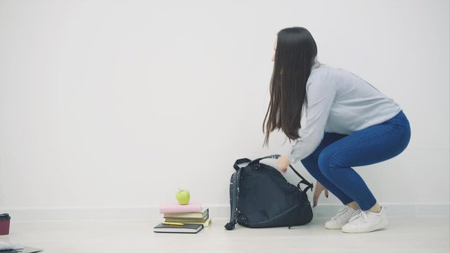 Adorable young asian girl appearing on white background, sitting down on the floor in lotos position, waving hello, openig up her backpack, taking out sketchbook.
