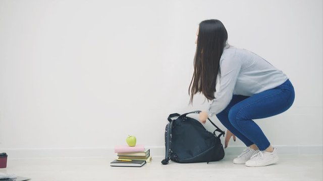 Adorable young asian girl appearing on white background, sitting down on the floor in lotos position, waving hello, openig up her backpack, taking out sketchbook.
