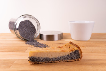 A piece of vegan cheesecake with poppy seeds together with a fork, a cup of coffee and poppy seeds on a wooden table