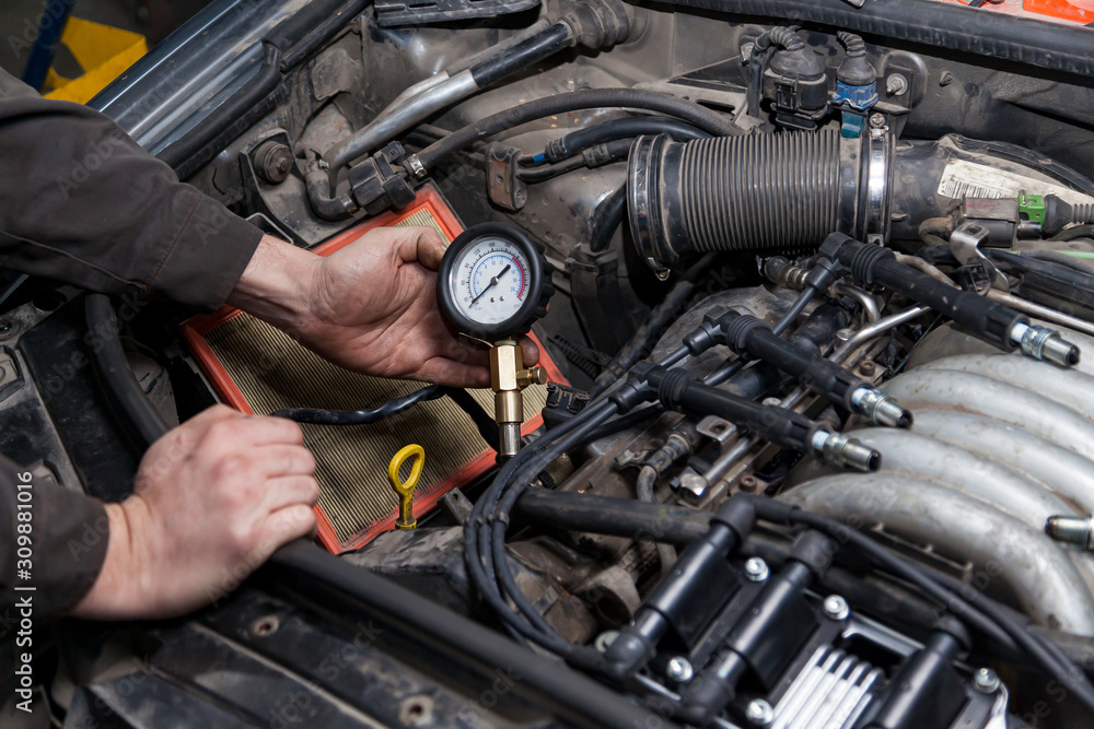 Fototapete A male mechanic measures the compression in the cylinder of a car engine using a barometer with a scale and an arrow during diagnosis and repair in a workshop for vehicles. Auto service industry.
