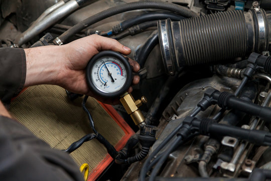 A male mechanic measures the compression in the cylinder of a car engine using a barometer with a scale and an arrow during diagnosis and repair in a workshop for vehicles. Auto service industry.